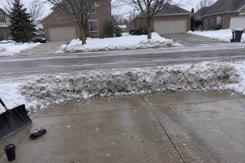 A ridge of dense plowed snow lying across the end of a concrete driveway where it meets the street, tire tracks in the road beyond it