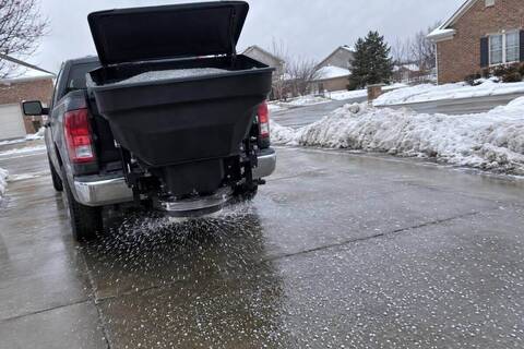 A tailgate spreader on the back of a pickup throwing granular ice melt across a wet driveway, pellets scattered on dark pavement