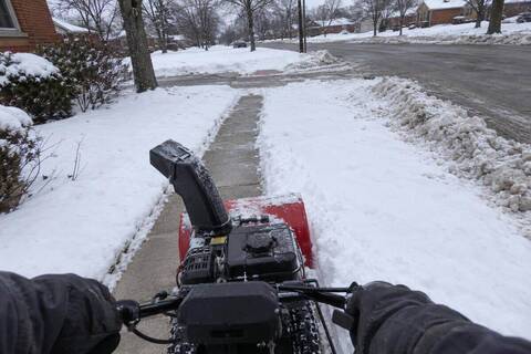 A walk-behind snowblower clearing a public sidewalk around a corner lot, chute turned away from the street, cut line in the snow behind