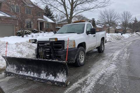 A pickup with a straight plow blade standing at a residential curb, blade resting on the pavement, snow-covered street behind